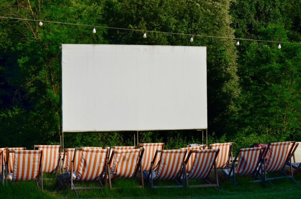 Empty outdoor cinema screen with striped deck chairs at dusk, surrounded by greenery