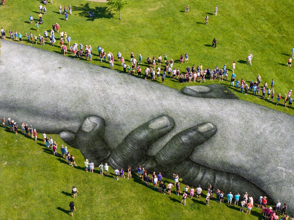 Aerial view of a giant painted fresco of intertwined hands on a lawn, with a long line of people holding hands along its edges