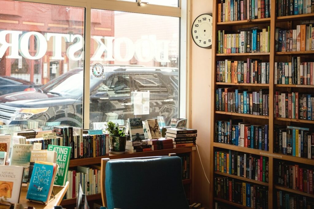 Corner of a small used bookstore with packed shelves, a book table and an armchair by the window
