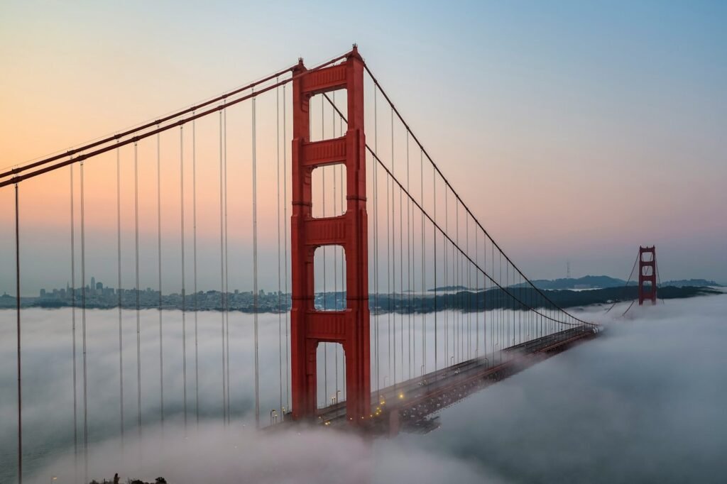 The Golden Gate Bridge's red tower rising above fog at sunrise, with the San Francisco skyline in the distance.
