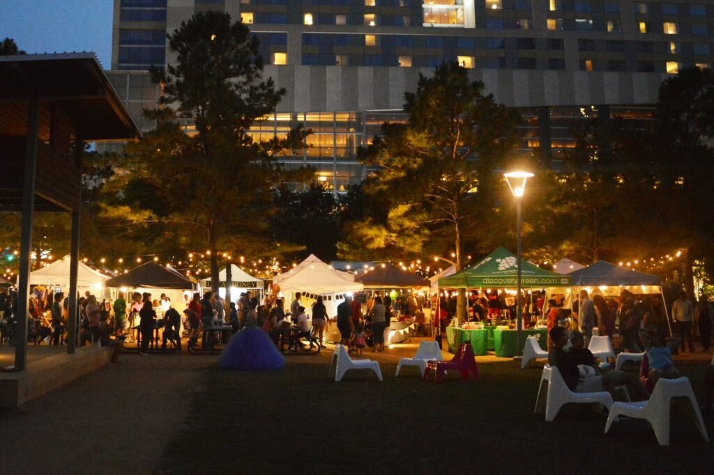 Crowds at an outdoor evening market of white tents under strings of lights in a city park.