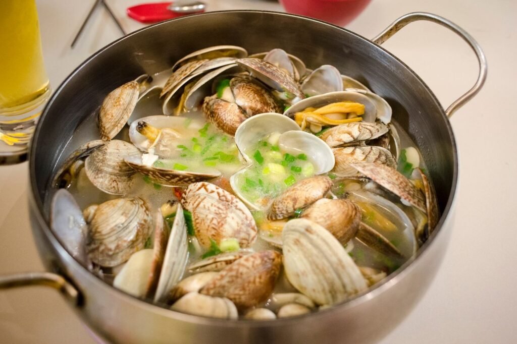 A steel pot of steaming clams in broth topped with chopped green onion.