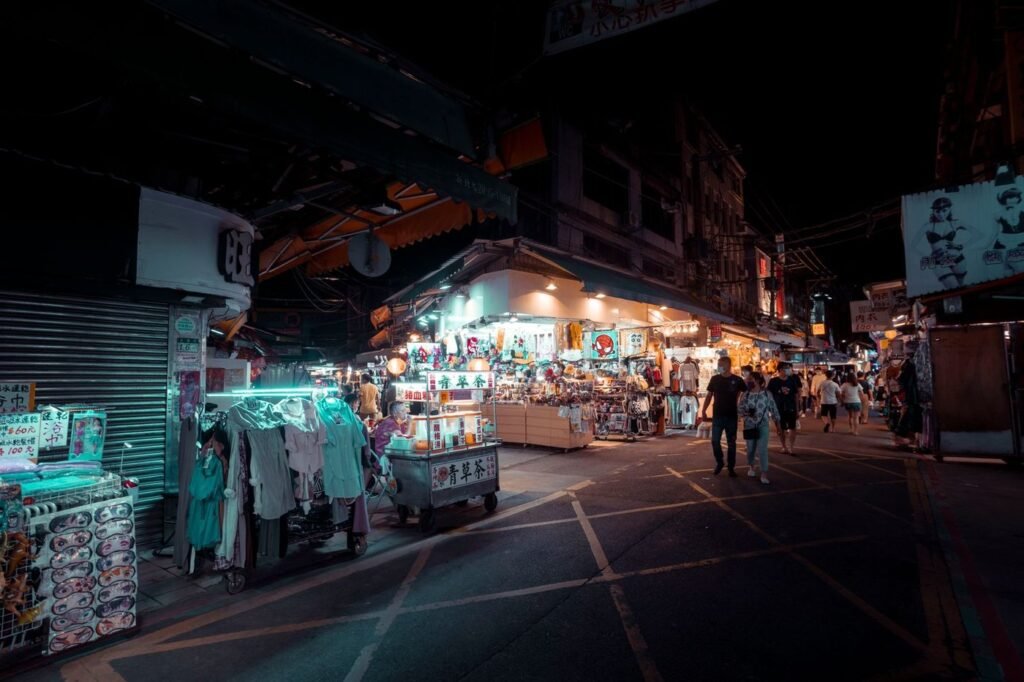 A lit night market street with food and clothing stalls and a few distant shoppers.