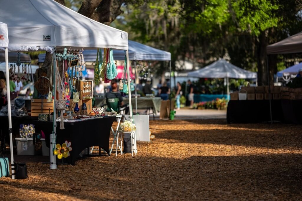 Vendor tents with hanging handmade goods on a shaded, sunlit outdoor market ground.