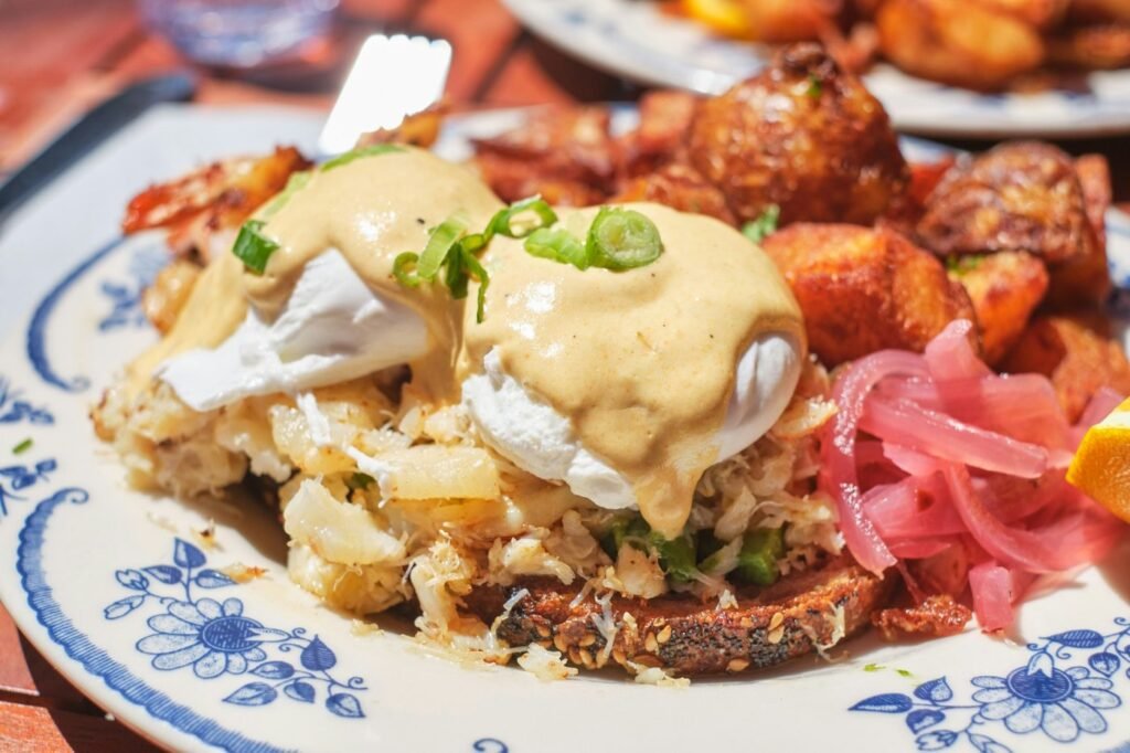 Poached eggs with hollandaise on crab cake with crispy potatoes and pickled onion on a blue-patterned plate.