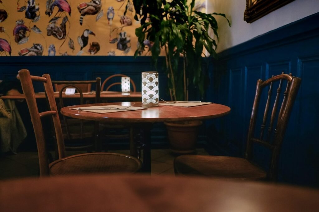 A small round wooden table set for two with a candle lamp in a warm, blue-panelled restaurant.