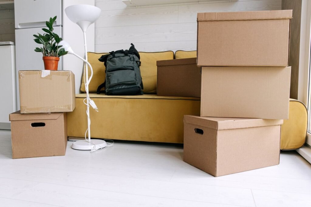Stacked cardboard moving boxes beside a yellow sofa with a backpack in a bright room.