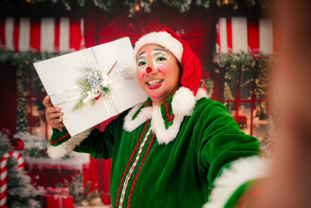 A smiling person in a green Christmas elf costume holding a gift box.