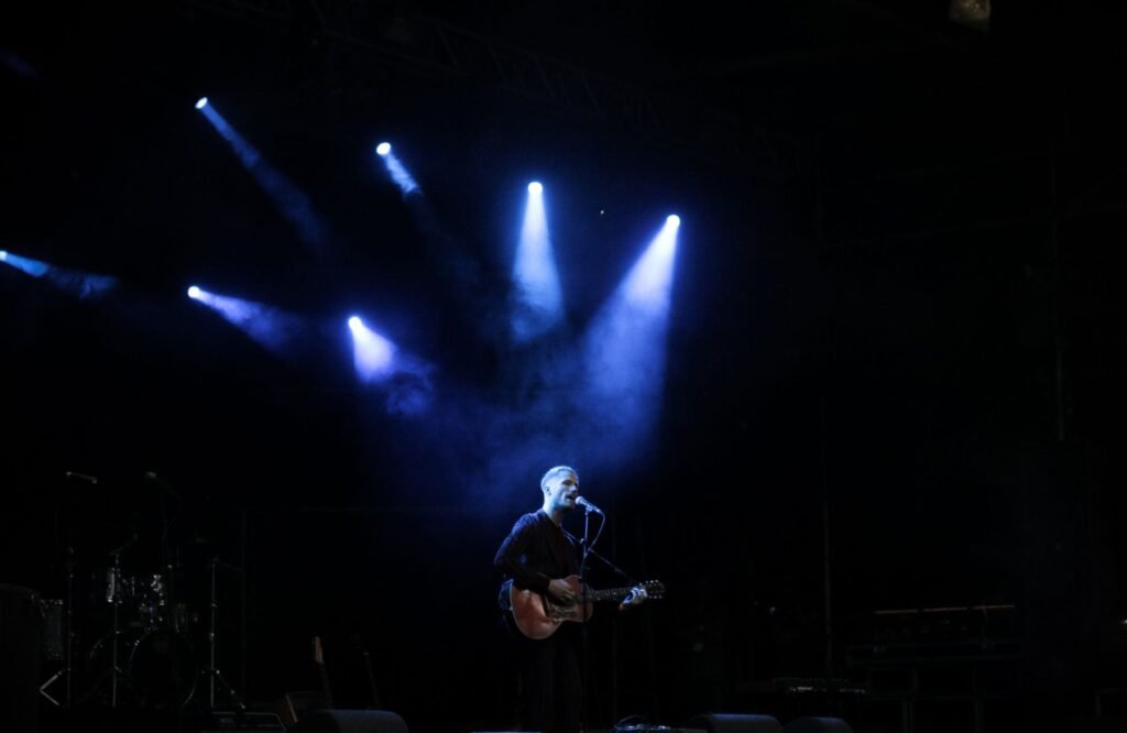 A solo musician with a guitar on stage under blue lights.