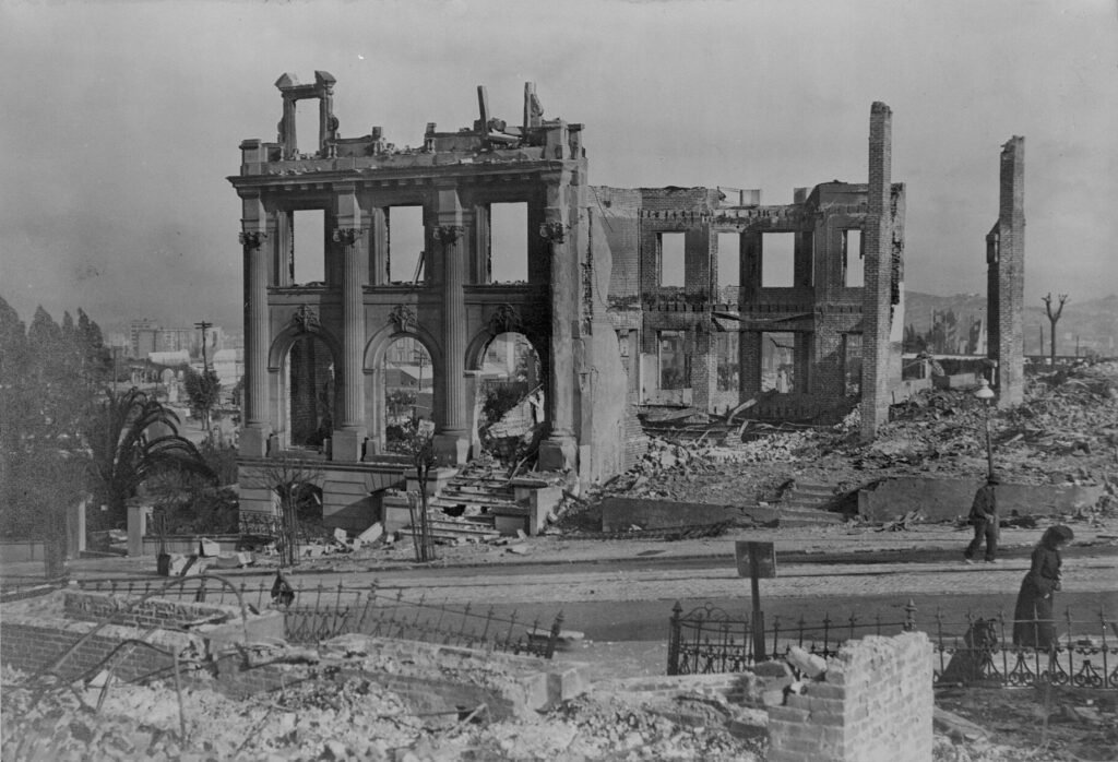 Burned shells of brick and stone buildings and rubble on a San Francisco street after the 1906 earthquake and fire, with two people walking past