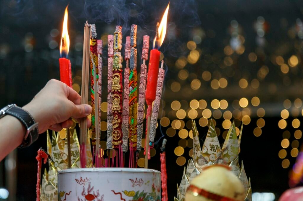 Incense sticks and red candles burning beside folded paper offerings at a Chinese altar.