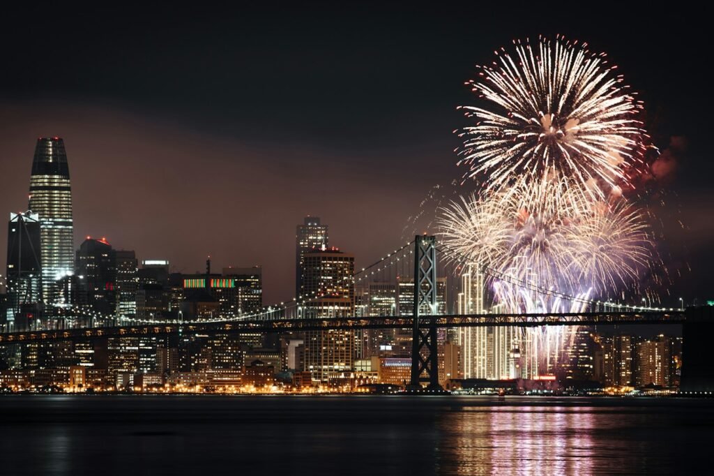 New Year's Eve fireworks bursting over San Francisco Bay at night, with the Bay Bridge and the downtown skyline including Salesforce Tower
