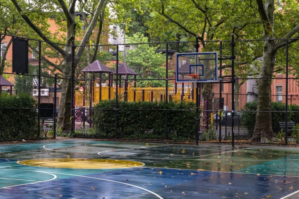Urban basketball court with a playground behind it among trees and buildings