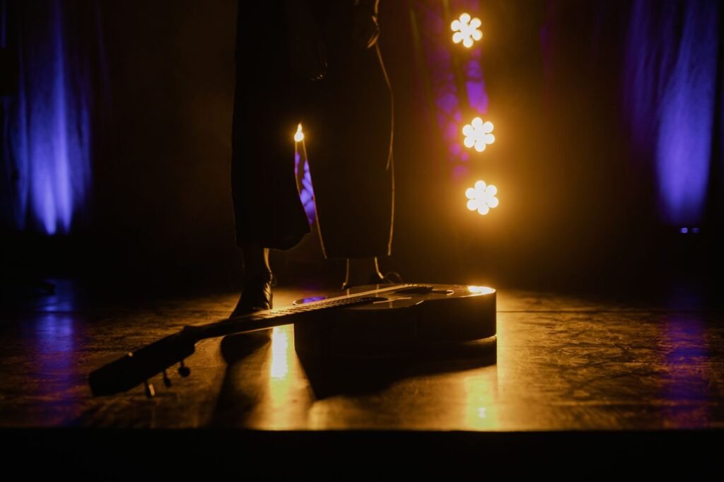 An acoustic guitar resting on a warmly lit theater stage, with a performer standing in shadow behind it